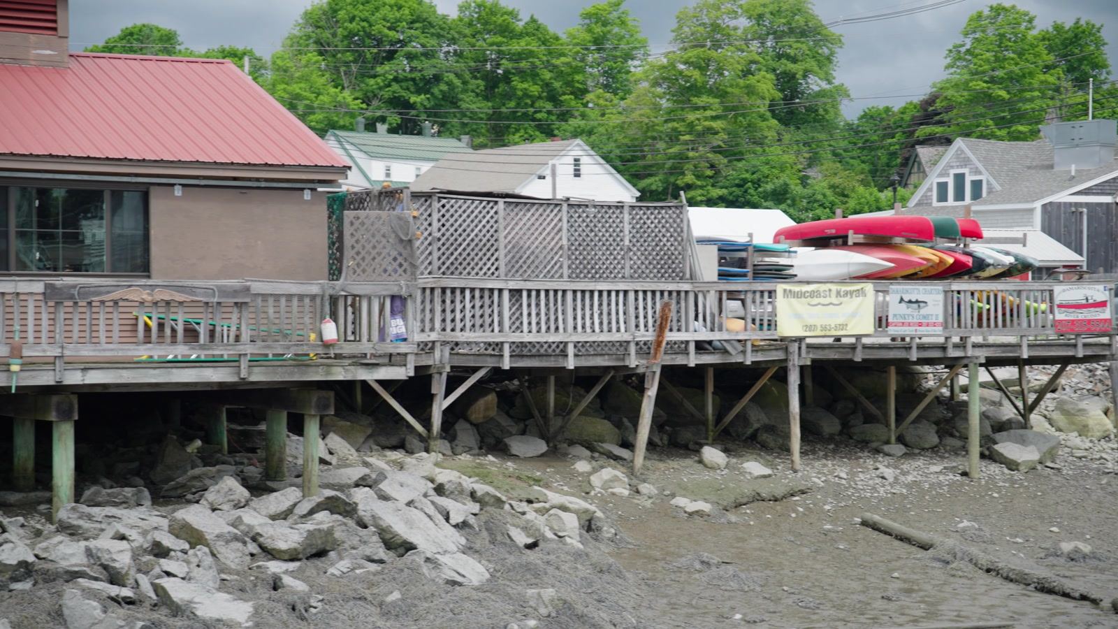 Buildings on pilings above the Damariscotta River at low tide, with kayaks on a deck